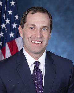 Jason Crow standing in front of an American flage wearing a dark suit, white button down shirt and tie while smiling