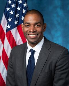 Antonio Delgado standing in front of an American flag wearing a dark suit, white button down shirt and navy blue tie while smiling