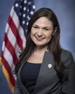 Abby Finkenauer standing in front of an American flag wearing a dark gray blazer and black shirt while smiling