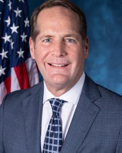 Harley Rouda stands in front of an American flag and wears a dark gray suit, white button down shirt and blue tie, half smiling
