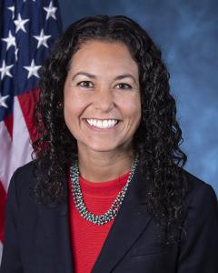 Xochitel Torres Small standing in front of an American flag wearing a dark blue blazer and red top while smiling