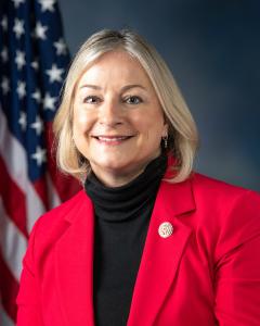 Susan Wild standing in front of an American flag wearing a red blazer and dark turtle neck shirt while smiling
