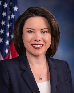 Angie Craig standing in front of an American flag wearing a dark blue blazer and smiling