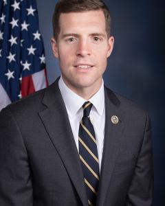 Conor Lamb standing in front of American flag while wearing a dark suit, white button down and striped tie