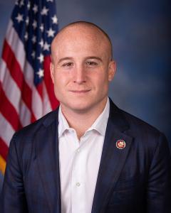 Max Rose standing in front of an American flag wearing a navy blue blazer and while button down, smiling