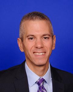 Anthony Brindisi standing in front of a blue background wearing a purple tie and shirt, black jacket