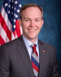 Ben McAdams standing in front of an American flag wearing a dark suit, light blue shirt and red tie