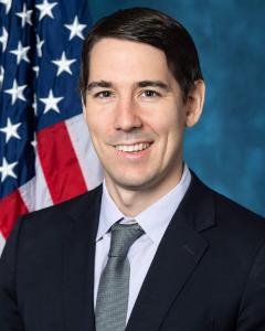 Josh Harder standing in front of an American flag wearing a dark suit and smiling