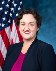 Katie Porter standing in front of the American flag wearing a dark blazer and magenta top while smiling
