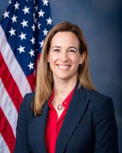 Mikie Sherrill standing in front of an American flag wearing a dark blazer and royal blue shirt while smiling