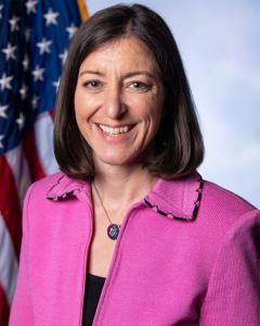 Elaine Luria standing in front of the American flag wearing a dark blazer and pearl necklace while smiling