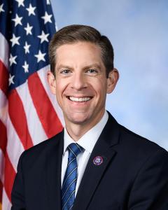Mike Levin standing in front of an American flag wearing a dark suit, blue tie and white button down and smiling