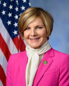 Susie Lee standing in front of an American flag wearing a pink jacket and white scarf and shirt