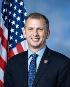 Sean Casten standing in front of an American flag wearing a dark blue suit, light blue button down shirt, blue striped tie and smiling