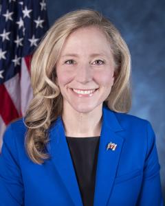 Abigail Spanberger standing in front of American flag, wearing bright blue blazer and dark shirt while smiling
