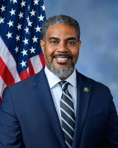 Steven Horsford standing in front of the American flag while wearing a dark suit, white button down shirt and blue tie, smiling