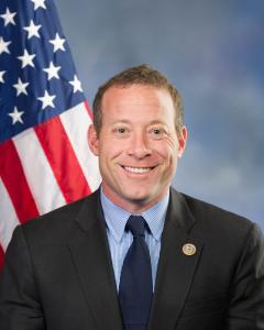 Josh Gottheimer standing in front of the American flag wearing a dark blue suit and tie withlighter blue button down and smiling